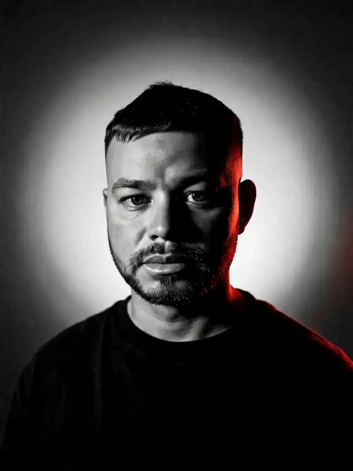 Black and white studio portrait of Jarryd Hennequin, half-lit against a radial glow with a single red rim light